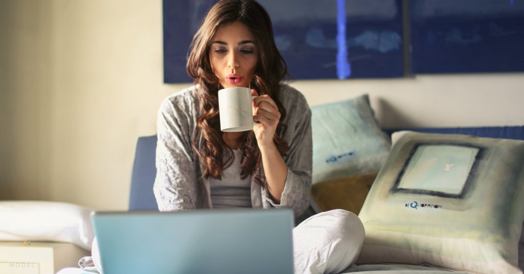 A woman researches breast augmentation cost on her laptop while taking a sip of coffee.