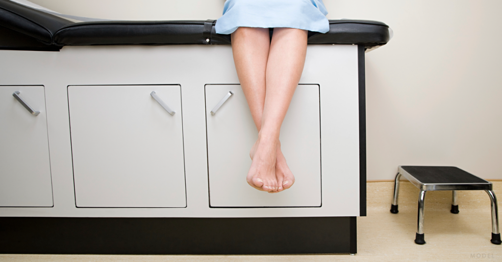 A woman sits on an exam table in preparation for her plastic surgery.