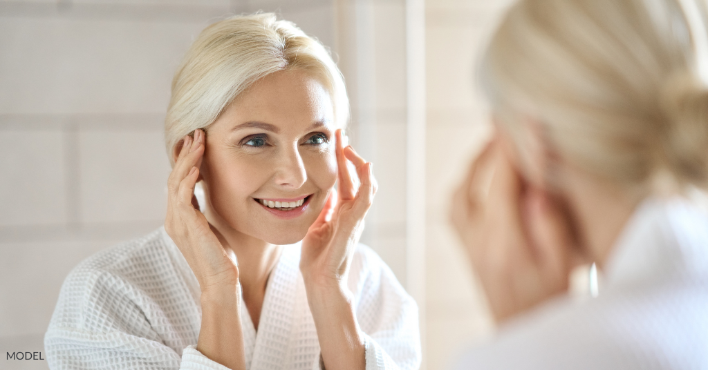 An older woman wearing a robe and looking at her face in the mirror (model)